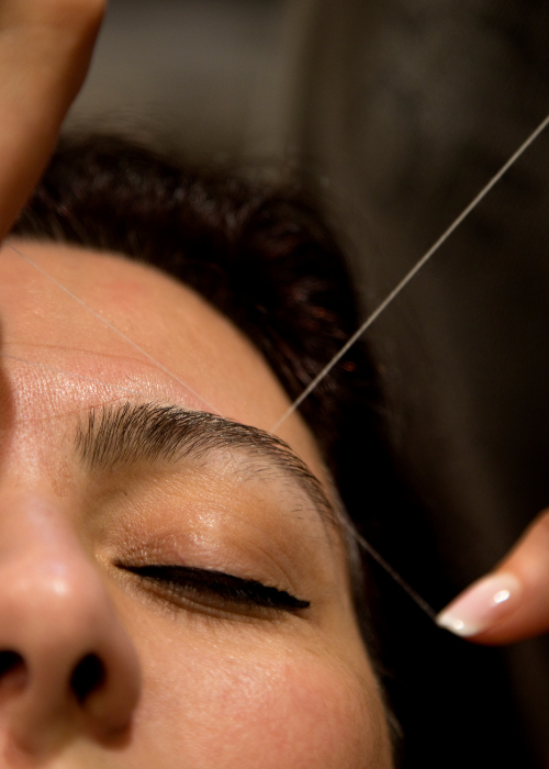 A woman sits in a salon chair as a beautician expertly threads her eyebrows, focusing on precision and detail.